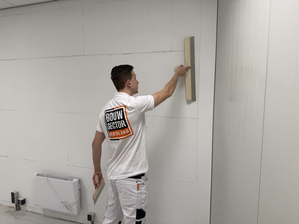 Stukadoor met Bouwsector-logo op zijn rug trekt een brede spaan omhoog over een wand van gipsblokken