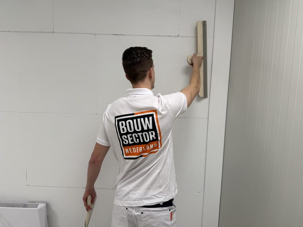 Stukadoor met het Bouwsector-logo werkt met een lange spaan het bovenste deel van een witte wand af
