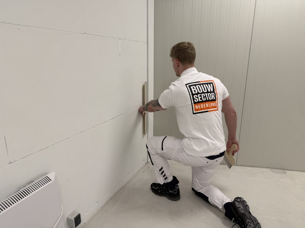 Stukadoor van Bouwsector Nederland zit op een knie en strijkt met een spaan de onderkant van een wand bij de plint