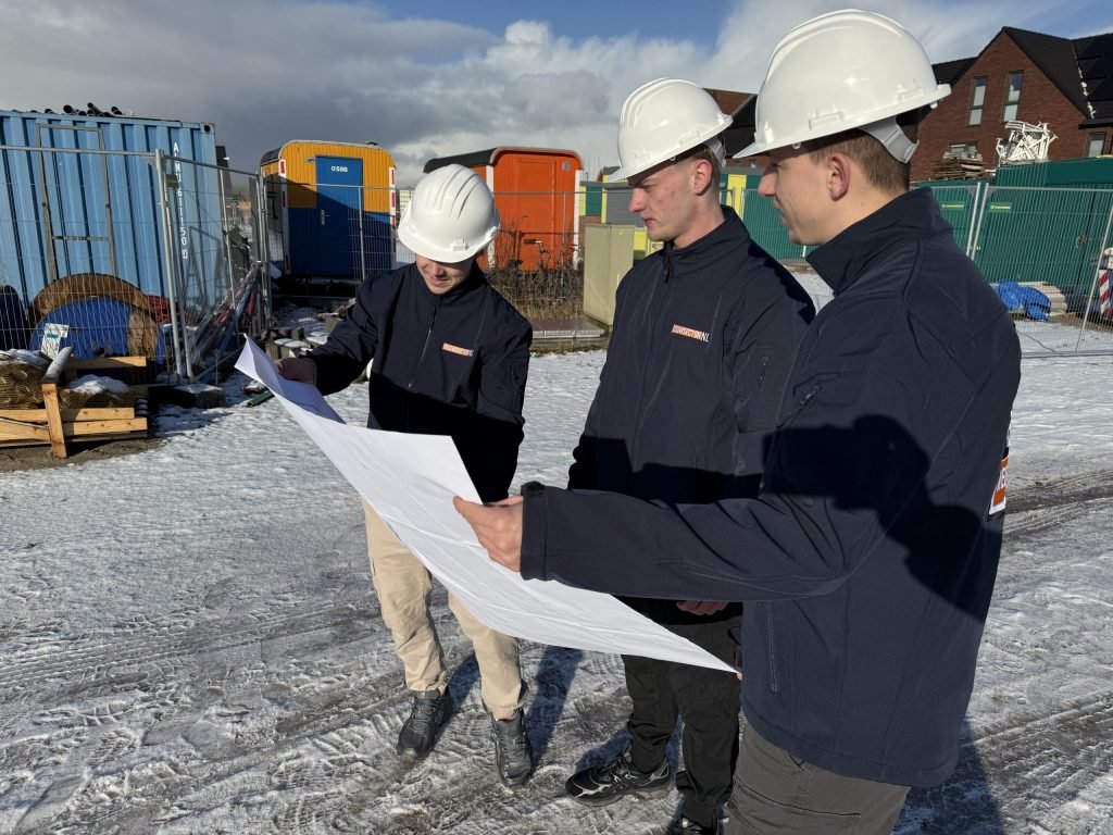 Drie vakmensen van Bouwsector Nederland met witte helm bespreken een bouwtekening op een nieuwbouwterrein
